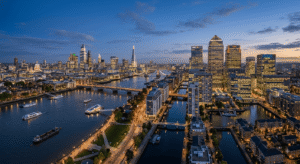 An aerial panoramic photograph at dusk capturing the illuminated skylines of both Canary Wharf (left, with One Canada Square) and the City of London (right, with The Shard, The Gherkin, and The Cheesegrater) separated by the River Thames in London, UK.